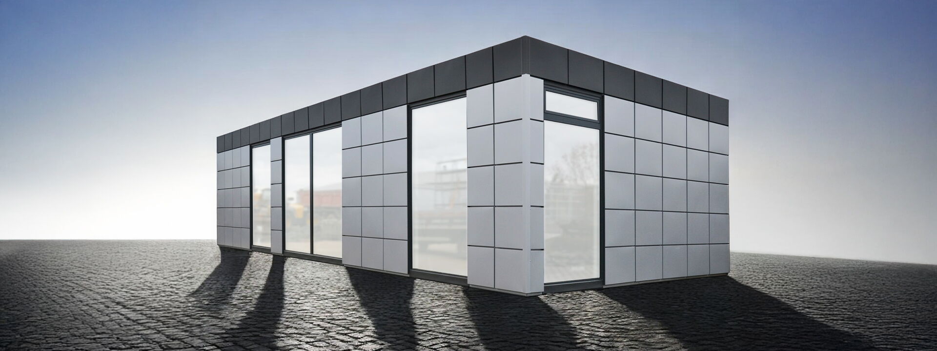Moderner Bürocontainer mit grauer Rasterfassade und großen bodentiefen Fenstern auf gepflasterter Fläche