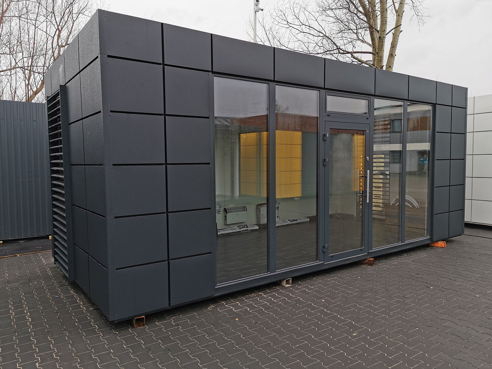 Moderner Bürocontainer mit dunkler Metallfassade und großflächiger Glasfront auf Pflasterfläche