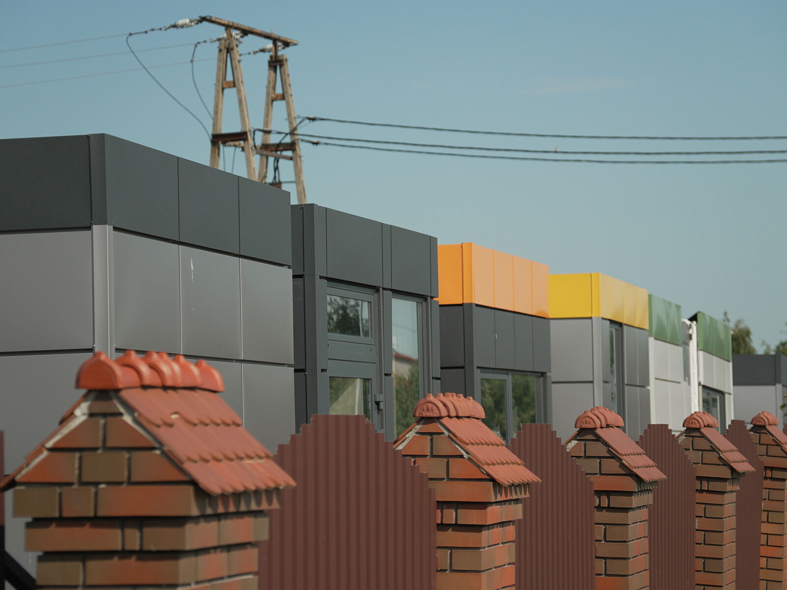 Reihe moderner Container mit farbigen Fassadenelementen hinter einem Ziegelzaun