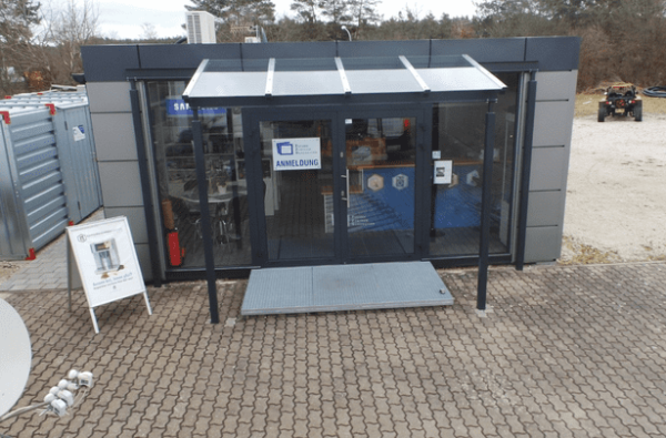 Eingangsbereich des Bürocontainers mit Glasvordach und Hinweisschild zur Anmeldung